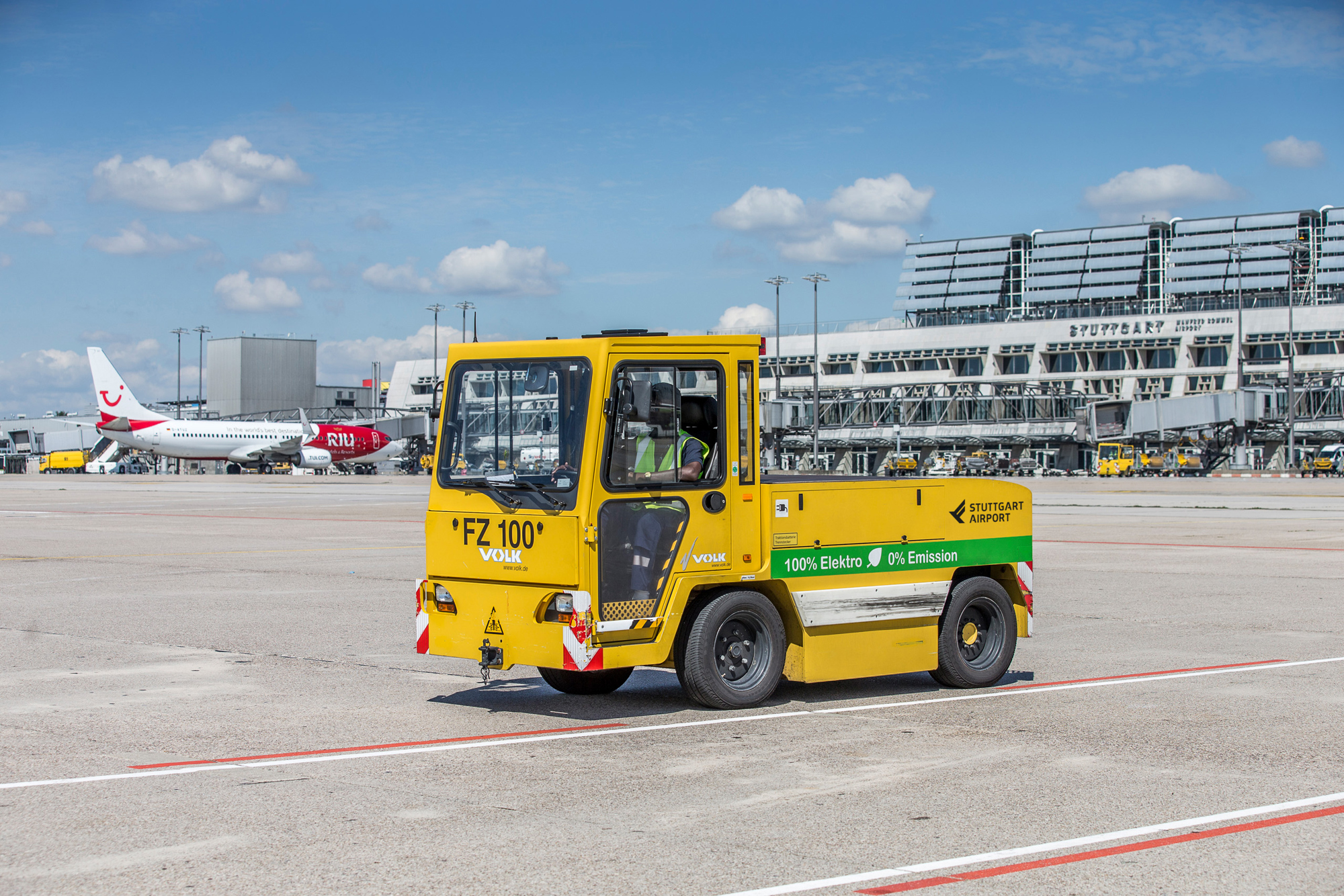 Ein elektrisch betriebener Gepäck-Schlepper steht auf dem Stuttgarter Flughafenvorfeld. Im Hintergrund stehen die Terminalgebäude. 