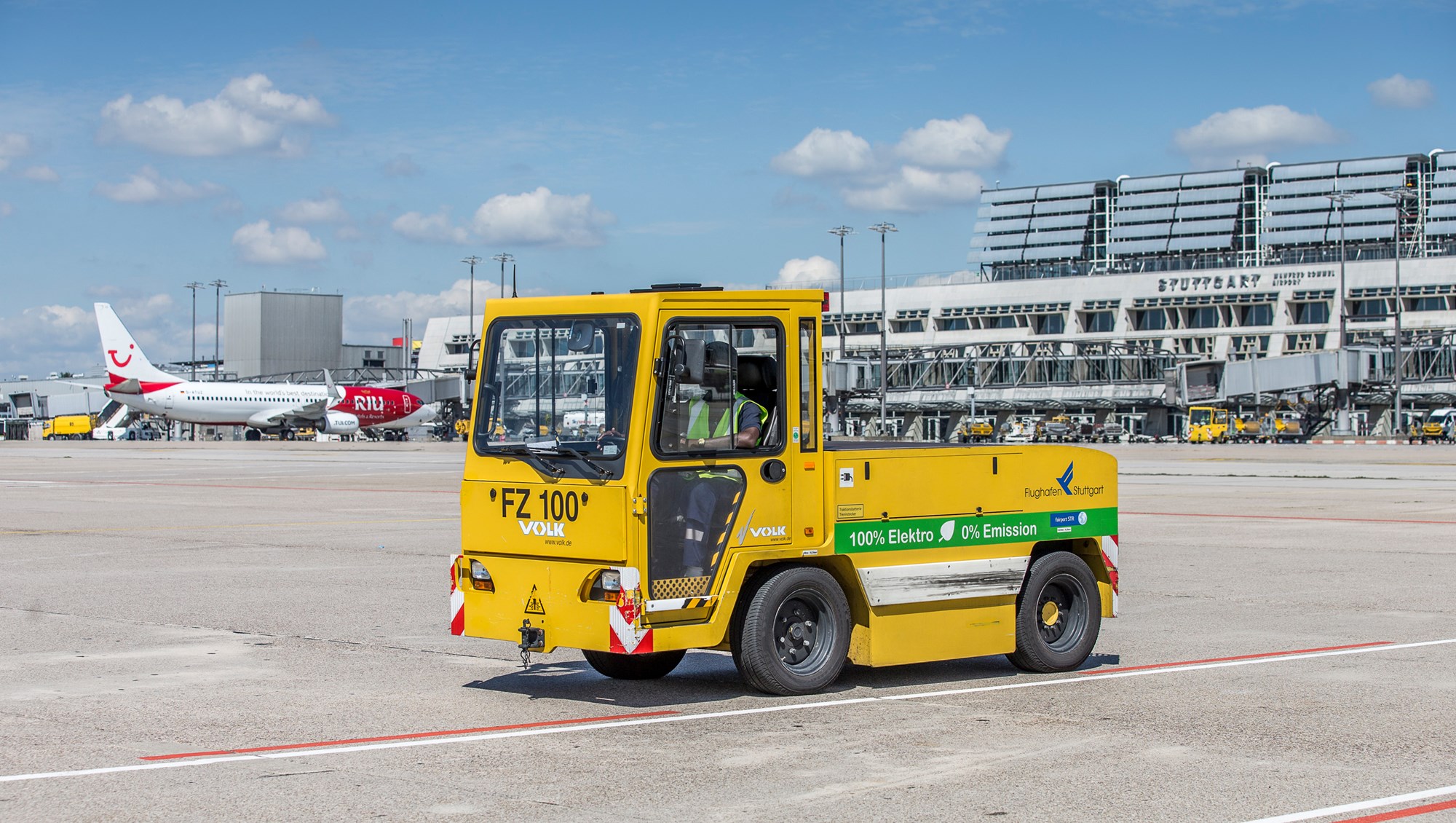 Ein elektrisch betriebener Gepäck-Schlepper steht auf dem Stuttgarter Flughafenvorfeld. Im Hintergrund stehen die Terminalgebäude. 