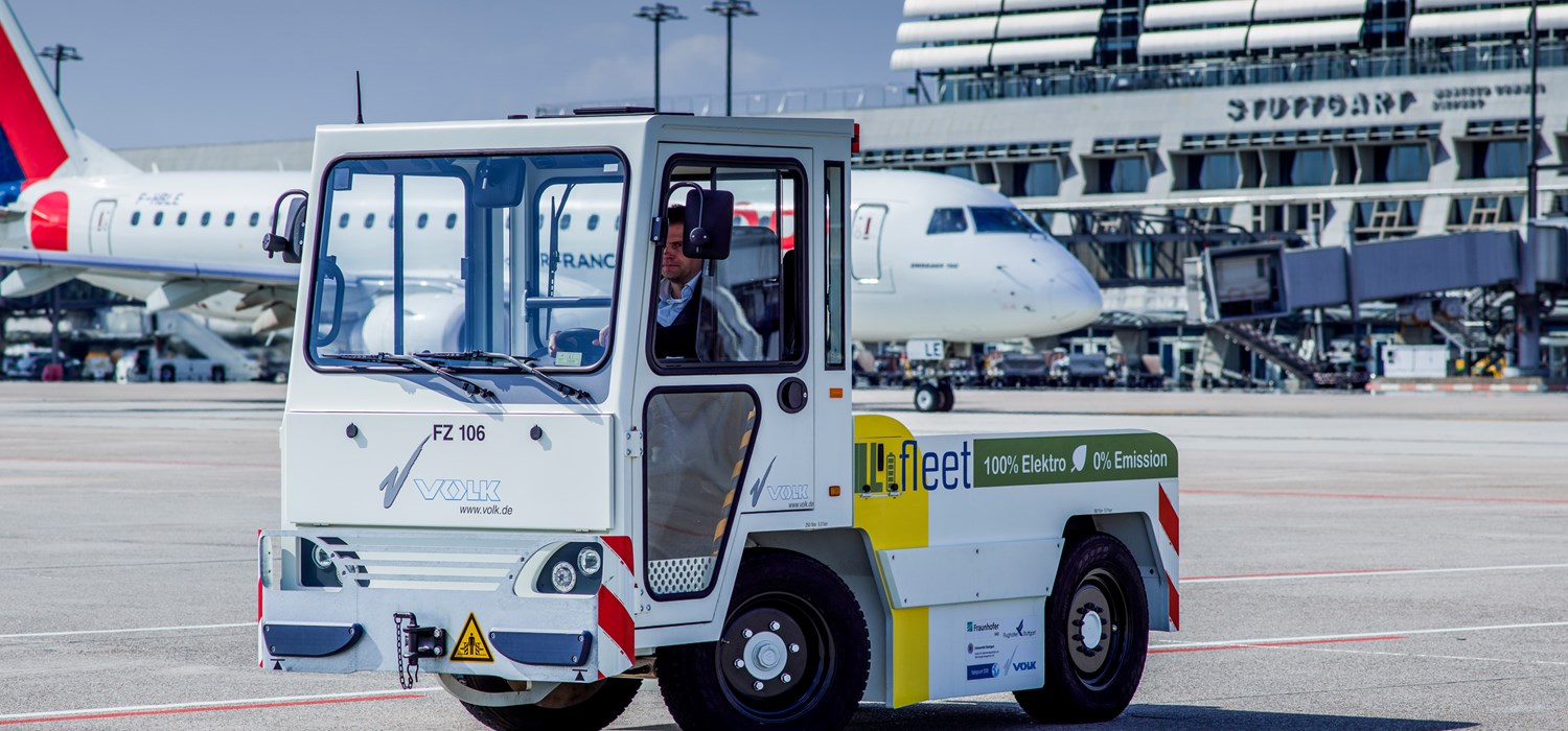 Ein elektrisch betriebener Gepäck-Schlepper mit Litium-Ionen-Batterie steht auf dem Stuttgarter Flughafenvorfeld. Im Hintergrund rollt ein Flugzeug zur Startposition. 