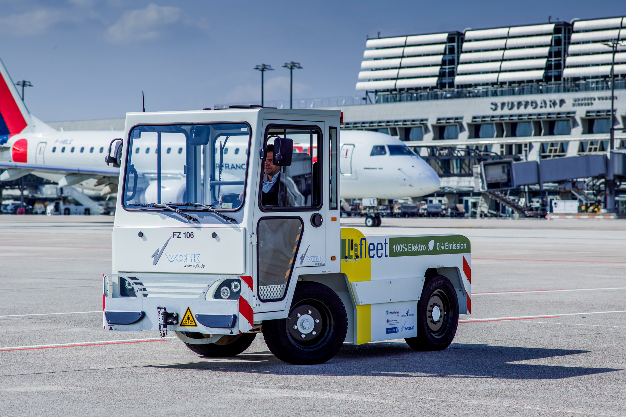 Ein elektrisch betriebener Gepäck-Schlepper mit Litium-Ionen-Batterie steht auf dem Stuttgarter Flughafenvorfeld. Im Hintergrund rollt ein Flugzeug zur Startposition. 