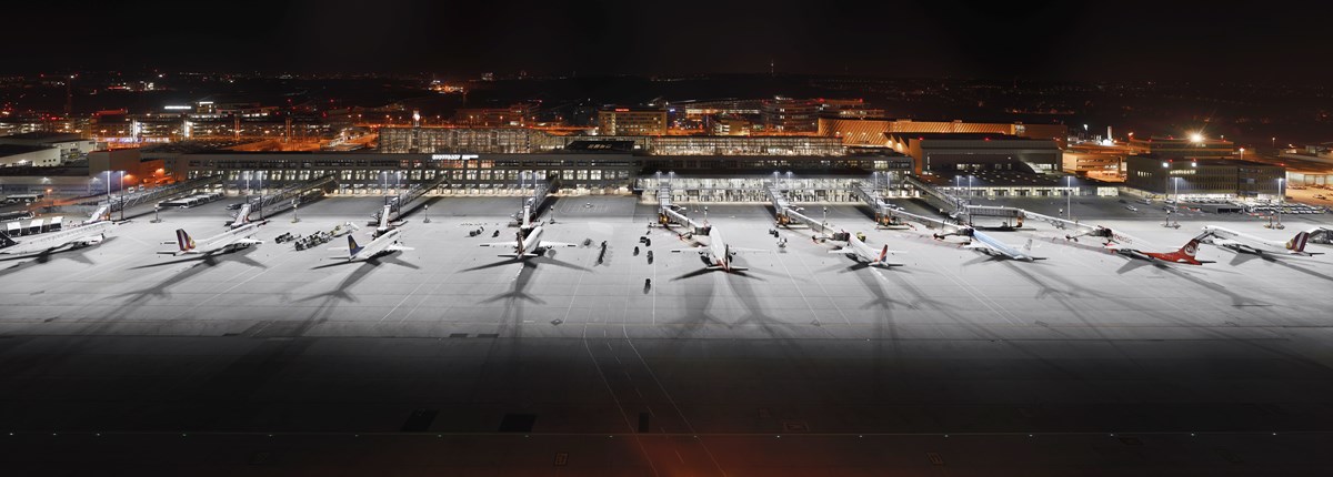 Vogelperspektive auf das beleuchtete Vorfeld und Terminalgebäude bei Nacht