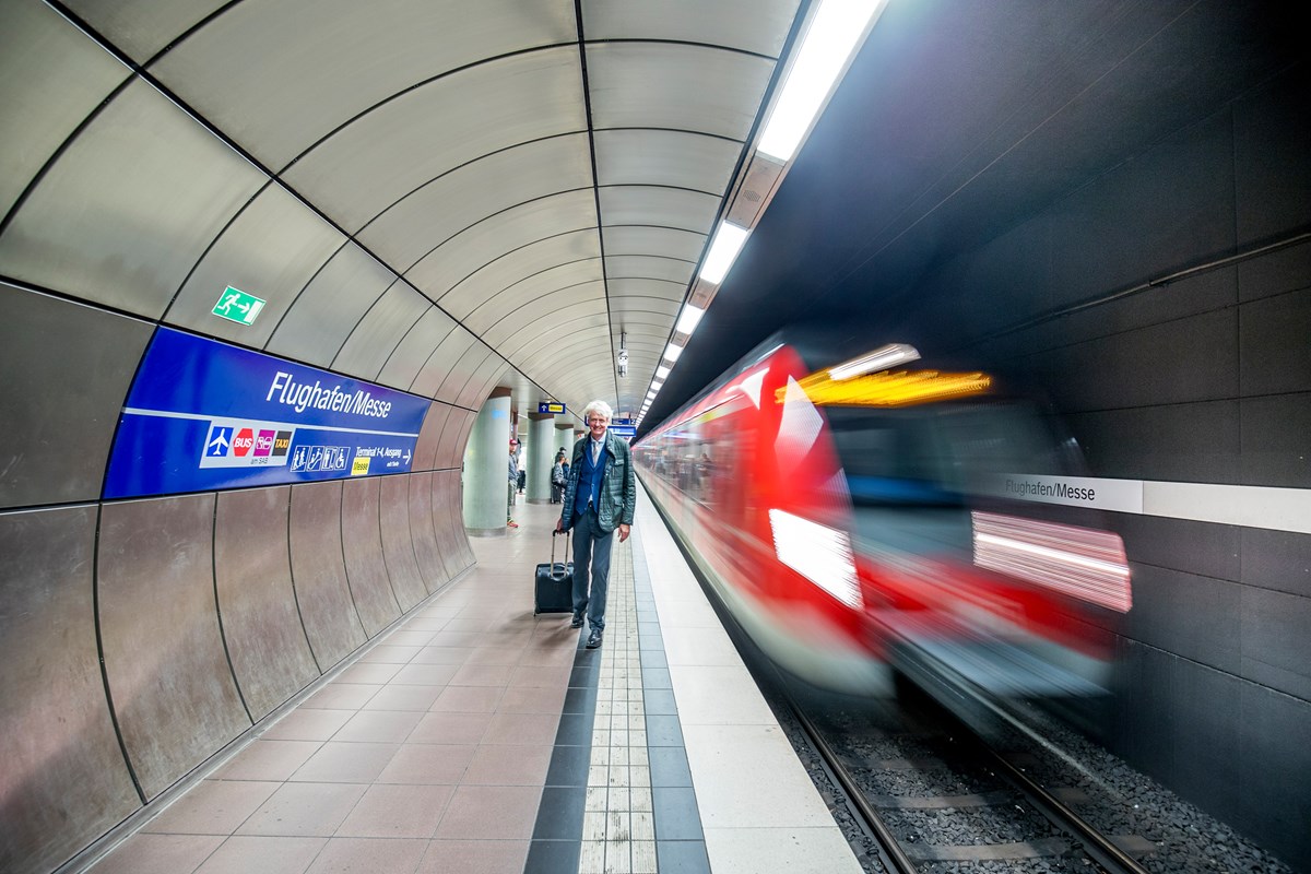 S-Bahn fährt an der Haltestellt Flughafen/Messe ein und Mann mit Koffer läuft auf dem Bahnsteig