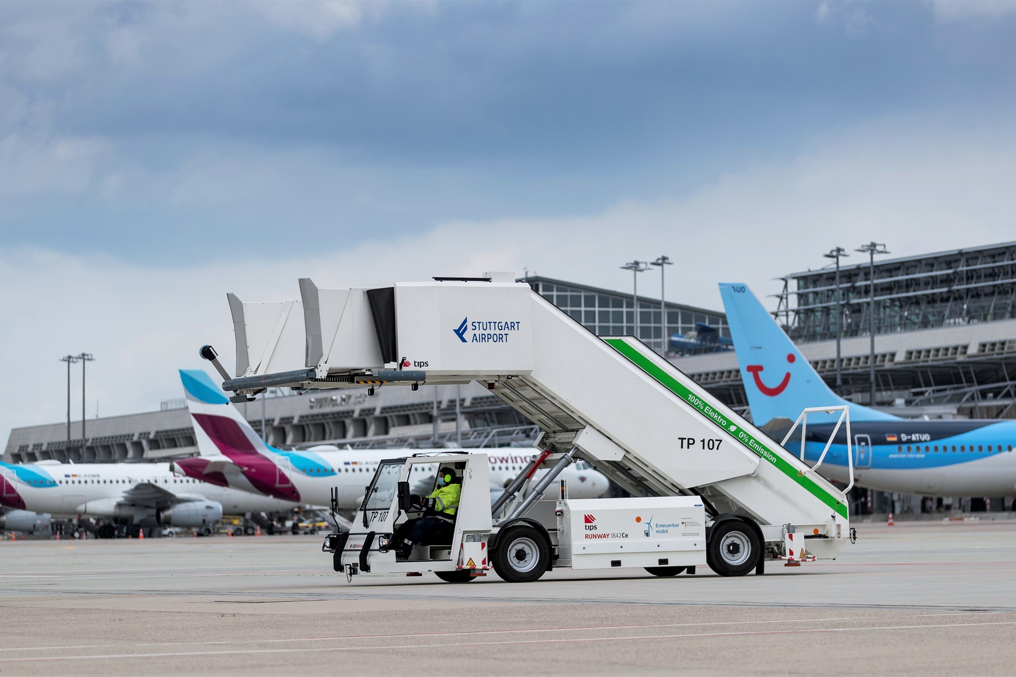 Eine elektrisch betriebene Passagiertreppe steht auf dem Stuttgarter Flughafenvorfeld. Im Hintergrund parken mehrere Flugzeuge. 