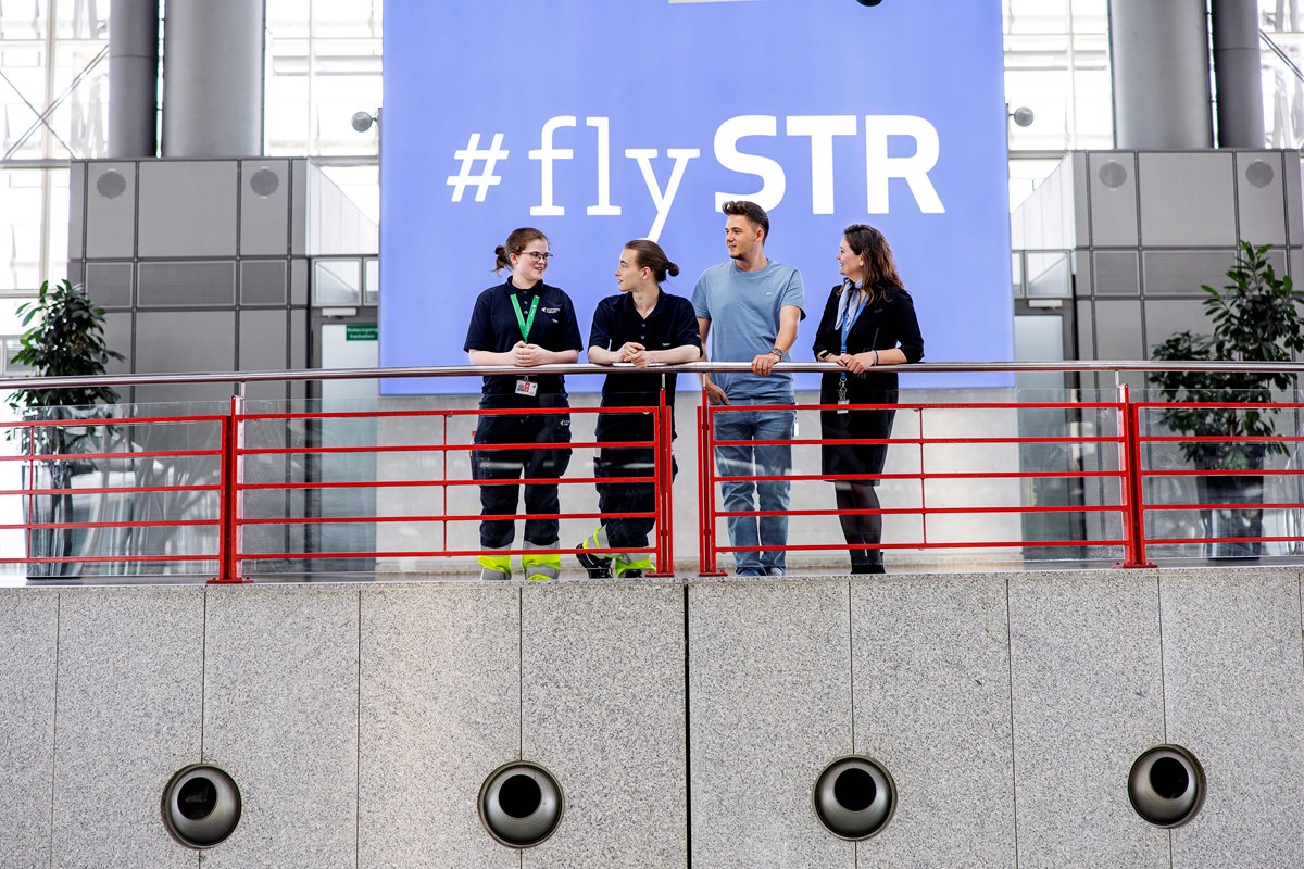 Zwei junge Frauen und zwei junge Männer in Flughafenkleidung lehnen sich an einem Geländer im Terminal des Flughafen Stuttgart an und sprechen miteinander. Hinter ihnen steht auf einer blauen Leuchtwand Hashtag fly STR. 