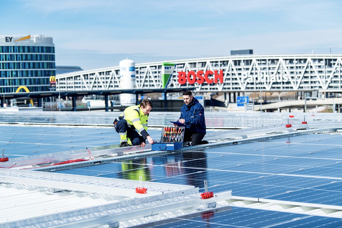 Zwei Mitarbeiter des Flughafen Stuttgart installieren eine Solaranlage auf einem Dach eines Flughafengebäudes.
