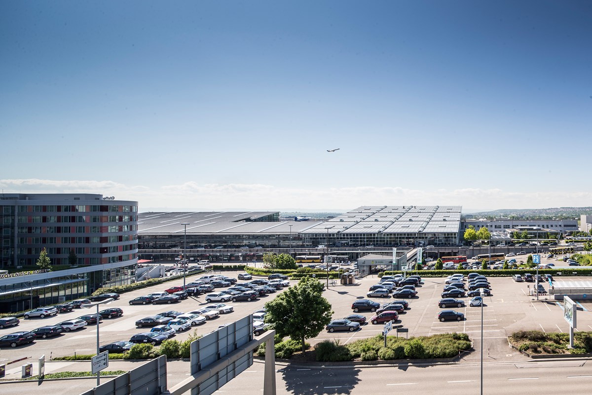 Blick auf den Parkplatz vor dem Terminalgebäude, im Hintergrund startet ein Flugzeug